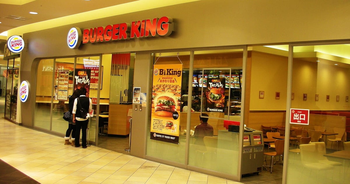 A Burger King restaurant in a shopping mall, with its red BURGER KING sign and round logos above a glass frontage, menu posters in the windows and two shoppers seen from behind.